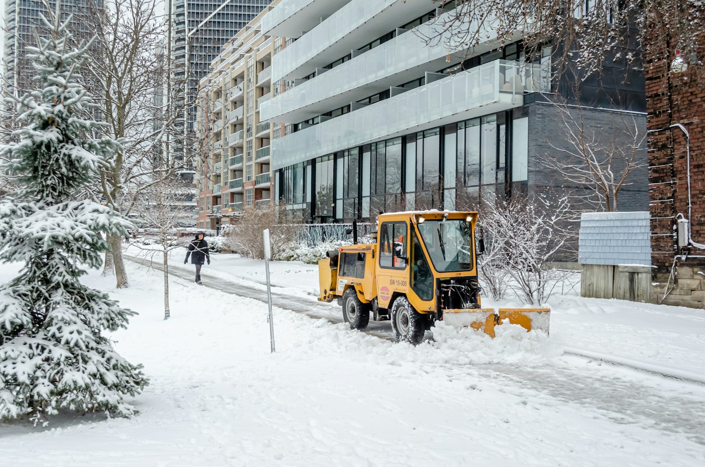 Yellow snowplow clearing a snowy street past apartment buildings.