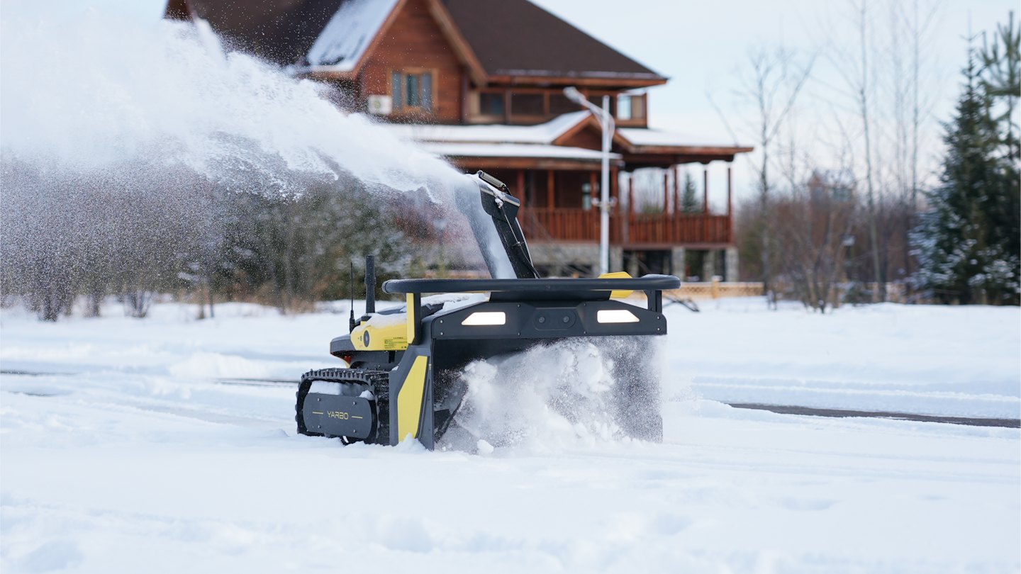 Yellow robotic snowblower clears snow from a paved residential driveway.